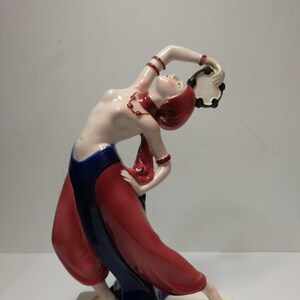 Może przedstawiać: Porcelanowa figurka tancerki w czerwono-niebieskim stroju, trzymającej tamburyn. Tancerka jest w dynamicznej, ekspresyjnej pozie.