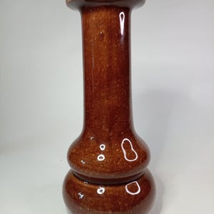 Può includere: Un vaso in ceramica marrone con finitura lucida. Il vaso ha una parte superiore ampia e piatta, un collo lungo e cilindrico e una base bulbosa. La base è composta da due sezioni arrotondate sovrapposte. Il vaso è alto circa 20 cm.