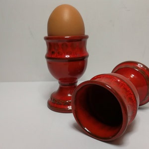 Può includere: Due porta uova in ceramica rossa, uno con un uovo marrone all'interno. I porta uova hanno una finitura testurizzata e smaltata.
