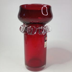 Puede incluir: Un jarrón de cristal rojo rubí con cuerpo cilíndrico y parte superior acampanada. La parte superior tiene forma redondeada con un anillo decorativo de cristal transparente alrededor del cuello. El jarrón tiene una etiqueta en el lateral.