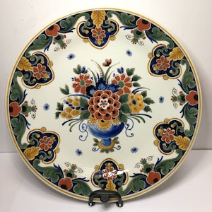 May include: A white ceramic plate with a blue, green, and gold floral design. The plate features a bouquet of red, orange, and yellow flowers in a blue vase. The plate has a gold rim and is decorated with a repeating pattern of stylized flowers and leaves.