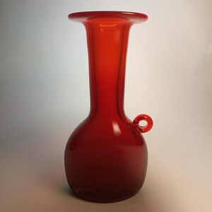 Könnte beinhalten: Eine rote Glasvase mit rundem Korpus und schmalem Hals. Die Vase hat einen kleinen Henkel an der Seite.
