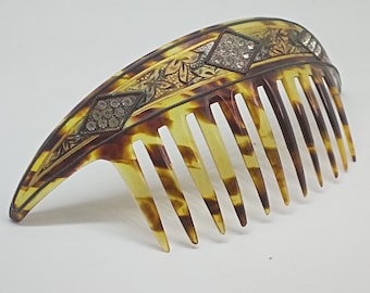 May include: An antique hair comb with a tortoiseshell design. The comb features a decorative silver band with diamond-shaped embellishments and floral detailing. The comb has long, evenly spaced teeth.