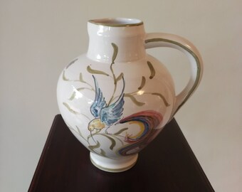 May include: A white ceramic pitcher with a green, blue, and red bird design. The pitcher has a wide base and a handle.