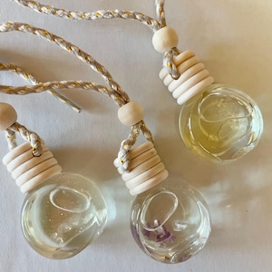 May include: Three round glass car air freshener bottles filled with clear liquid and small crystals. Each bottle features a light-coloured wooden cap, a wooden bead, and a braided white cord with gold metallic threading for hanging purposes.