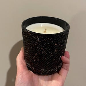 Könnte beinhalten: Eine schwarze Kerze in einem strukturierten Glasbehälter. Die Kerze hat weißes Wachs und einen einzelnen Docht. Der Behälter hat einen schwarzen Rand und ein Muster aus kleinen, kupferfarbenen Flecken. Die Kerze wird in einer Hand gehalten.
