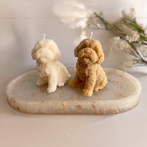 May include: Two small dog-shaped candles on a white oval tray with gold flecks. One candle is white and the other is brown.