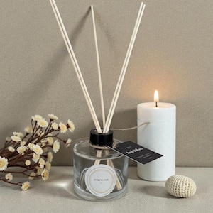 May include: A clear glass reed diffuser with wooden reeds, a black cap, and a label that reads "FORTH OAK". A lit white candle and a small decorative item are next to the diffuser. The background is a neutral colour.