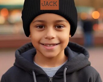 May include: A black knit beanie with a brown leather patch that says "JACK" on it. The beanie is being worn by a young person with a black hooded sweatshirt.