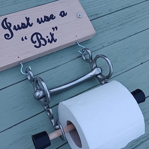 May include: A wooden sign with the text "Just use a "Bit" hanging on a light blue wooden wall. A toilet paper roll is attached to a silver metal horse bit.