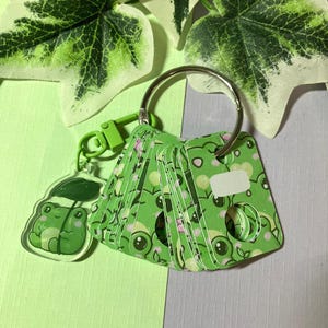 Può includere: Un portachiavi a tema rana verde con un ciondolo in acrilico trasparente e una serie di piccole carte decorate. Le carte presentano un motivo a rana ripetuto e sono attaccate a un anello d'argento.