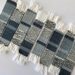 May include: A set of seven woven bookmarks with a white fringe. The bookmarks are made with a blue and white yarn and have a geometric pattern.