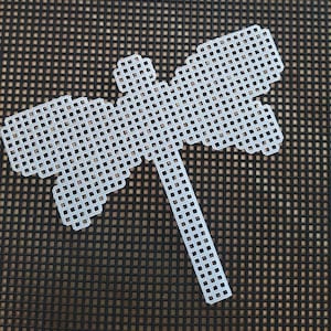 May include: White plastic dragonfly pattern on a black gridded surface. The pattern is made up of small squares.