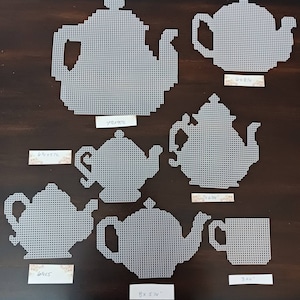 May include: Seven white teapots and a white teacup made of plastic canvas. The teapots are different sizes and shapes. The teacup is 3 inches tall. The teapots are labeled with their dimensions in inches.