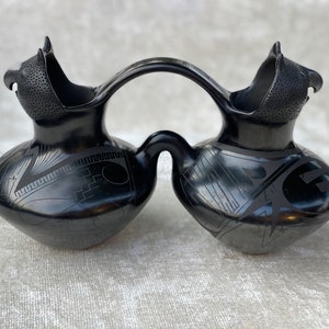 May include: Black ceramic double-spouted vase with a bridge-like handle. The vase is decorated with geometric patterns and has a bird-like spout on each side.