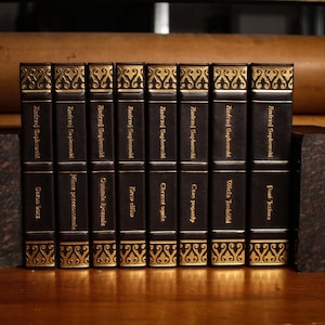 May include: A set of ten black hardback books with gold accents and titles in Polish. The books are arranged vertically and are all by the same author, Andrzej Sapkowski. The titles of the books are: "Sezon burz", "Miecz przeznaczenia", "Ostatnie życzenie", "Krew elfów", "Chrzest ognia", "Czas pogardy", "Wieża Jaskółki", "Pani Jeziora".