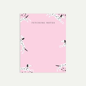May include: A pink notepad with a playful design featuring white dalmatian dogs with black spots and hearts. The words "FETCHING NOTES" are printed in the centre. The notepad is rectangular and sits on a white background.