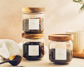 May include: Three glass jars with wooden lids filled with loose leaf tea. The jars are labeled "Fruit Tea", "Chai Tea", and "Earl Grey Tea".