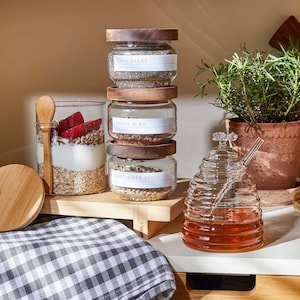 May include: Three glass jars with wooden lids filled with chia seeds, cacao nibs, and sunflower seeds. The jars are stacked on top of each other. A clear glass honey dispenser shaped like a beehive with a straw is on a white surface.