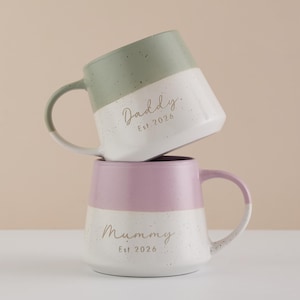 May include: Two ceramic mugs, one stacked on top of the other. The top mug is green and white, with the word "Daddy" and "Est 2026" printed on it. The bottom mug is pink and white, with the word "Mummy" and "Est 2026" printed on it.