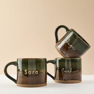 May include: Three ceramic mugs with a dark green and brown glaze are stacked. The mugs have a rounded shape with a handle and the names "Sara", "Gigi", and "Sarah" are written on them in white script.
