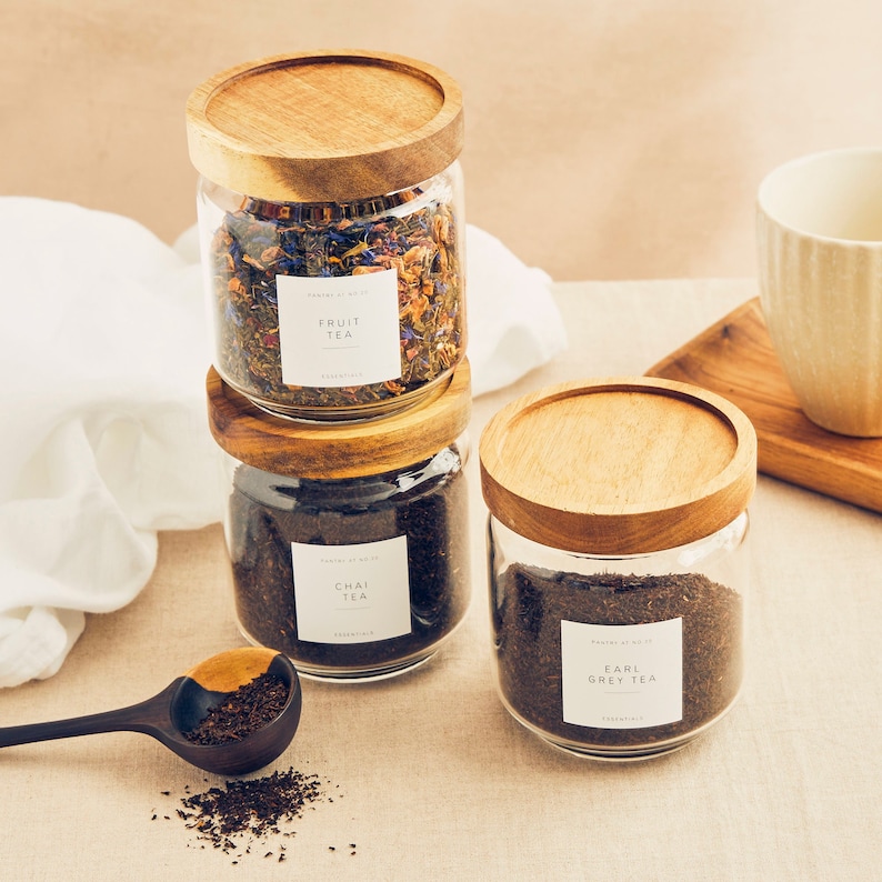 May include: Three glass jars with wooden lids filled with loose leaf tea. The jars are labeled "Fruit Tea", "Chai Tea", and "Earl Grey Tea".