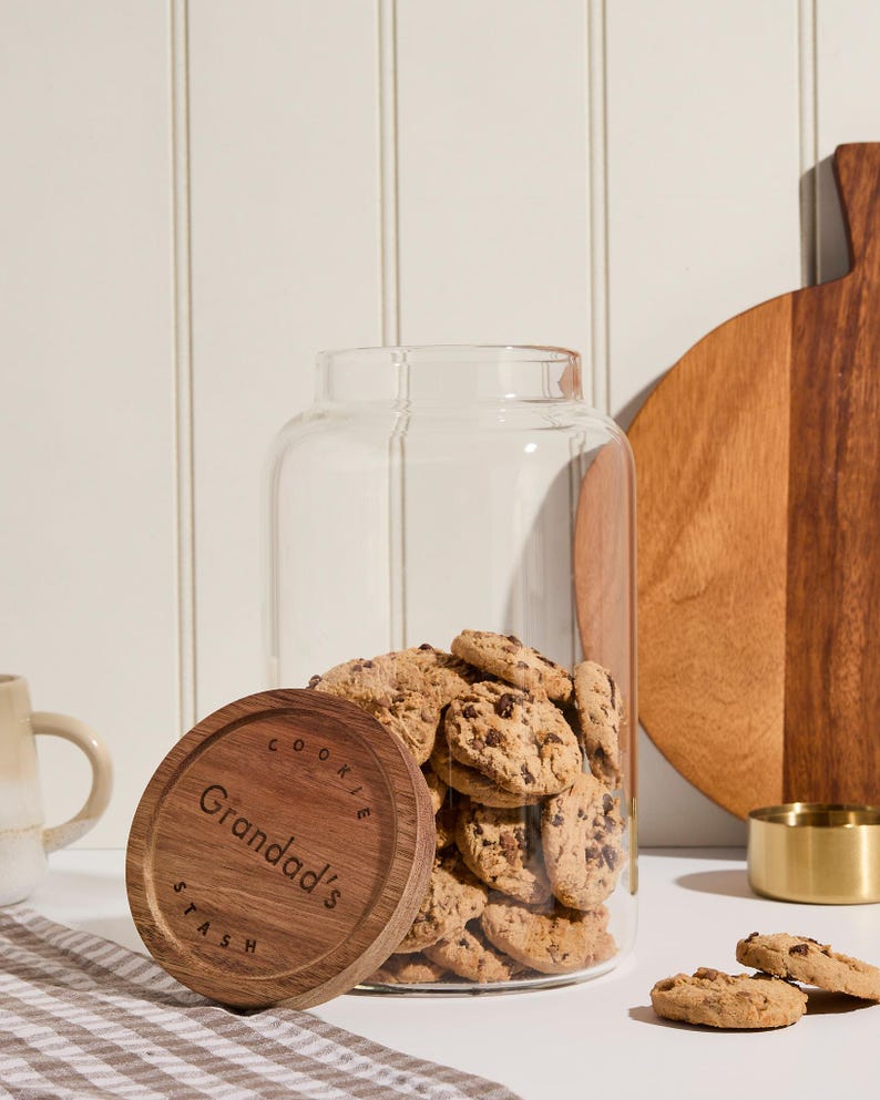 Peut inclure: Un pot &agrave; biscuits en verre avec un couvercle en bois. Le couvercle porte la gravure "Cookie Stash" et "Granddad's". Le pot est rempli de biscuits aux p&eacute;pites de chocolat.