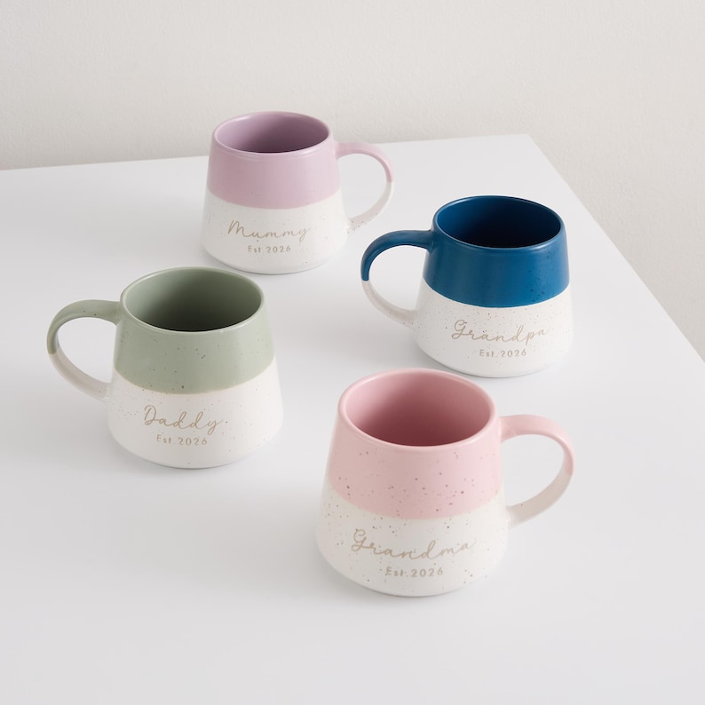 May include: Four ceramic mugs in pastel colors: pink, blue, green, and pink. Each mug has a white speckled base and a colored top. The mugs are labeled "Mummy", "Grandpa", "Daddy", and "Grandma", with "Est 2026" below each name.