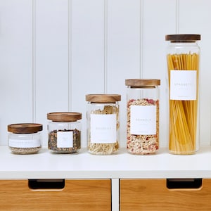 May include: Five glass jars with wooden lids and labels on a white shelf. The labels read "Sunflower Seeds", "Small Tea", "Noodles", "Granola", and "Spaghetti".