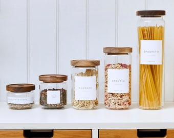 May include: Five glass jars with wooden lids and labels on a white shelf. The labels read "Sunflower Seeds", "Small Tea", "Noodles", "Granola", and "Spaghetti".