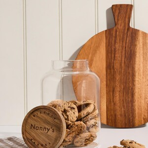 Peut inclure: Un bocal en verre avec un couvercle en bois sur lequel est inscrit "Biscuit Barrel Nanny's" rempli de biscuits aux p&eacute;pites de chocolat. Le bocal est pos&eacute; sur une surface blanche avec un tissu &agrave; carreaux brun et blanc.