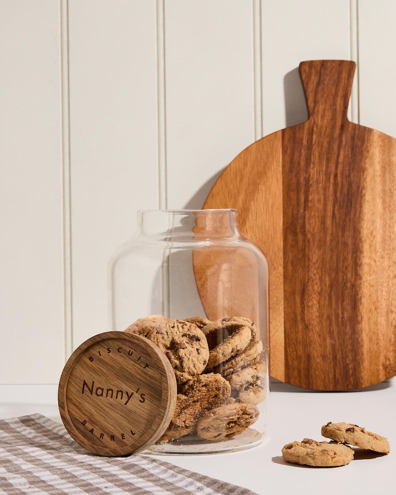 Peut inclure: Un bocal en verre avec un couvercle en bois sur lequel est inscrit "Biscuit Barrel Nanny's" rempli de biscuits aux p&eacute;pites de chocolat. Le bocal est pos&eacute; sur une surface blanche avec un tissu &agrave; carreaux brun et blanc.