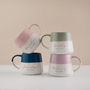 May include: Four ceramic mugs stacked, each with a different color scheme and text. The mugs are white with a speckled finish and a colored top half. The text on the mugs reads "Grandma", "Daddy", "Grandpa", and "Mummy", with "Est. 2026" below each.