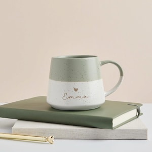 May include: A two-toned ceramic mug with a speckled finish, featuring a sage green upper section and a white lower section. The white portion is printed with the name Emma in cursive script, accompanied by a small heart icon above the text.