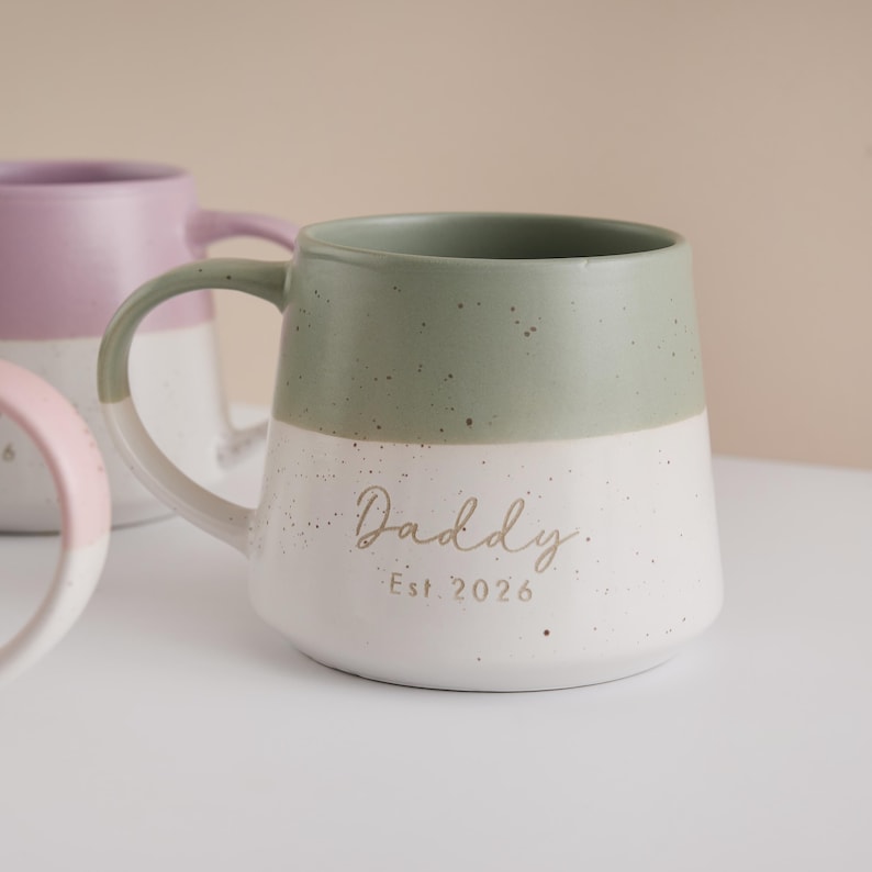 May include: A ceramic mug with a green top half and a white bottom half. The mug has a curved handle and the word "Daddy" and "Est. 2026" printed in gold lettering. Another mug is visible in the background.