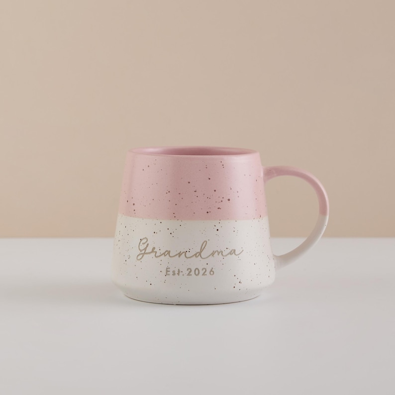 May include: A two-tone ceramic mug with a pink top and white speckled bottom. The word "Grandma" is written in cursive, with "Est. 2026" below it. The mug has a curved handle and is set against a neutral background.