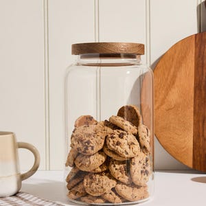 Peut inclure: Un bocal en verre transparent avec un couvercle en bois rempli de biscuits aux p&eacute;pites de chocolat. Le bocal est pos&eacute; sur un comptoir blanc avec une serviette de cuisine &agrave; carreaux.