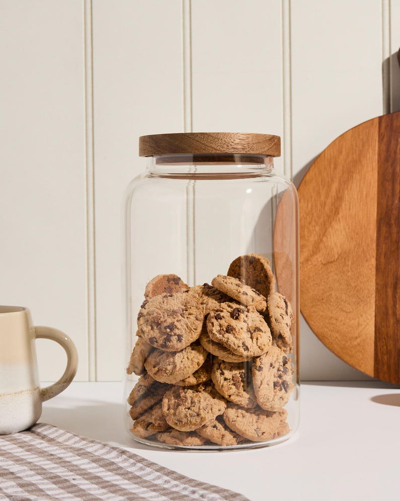 Peut inclure: Un bocal en verre transparent avec un couvercle en bois rempli de biscuits aux p&eacute;pites de chocolat. Le bocal est pos&eacute; sur un comptoir blanc avec une serviette de cuisine &agrave; carreaux.