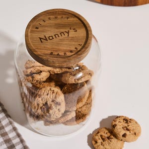 Peut inclure: Un bocal en verre avec un couvercle en bois sur lequel est inscrit "Biscuit Barrel Nanny's" est rempli de biscuits aux p&eacute;pites de chocolat. Deux biscuits sont pos&eacute;s sur une surface blanche &agrave; c&ocirc;t&eacute; du bocal.