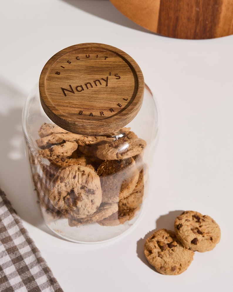Peut inclure: Un bocal en verre avec un couvercle en bois sur lequel est inscrit "Biscuit Barrel Nanny's" est rempli de biscuits aux p&eacute;pites de chocolat. Deux biscuits sont pos&eacute;s sur une surface blanche &agrave; c&ocirc;t&eacute; du bocal.