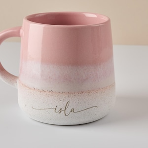 May include: A pink and white ceramic mug with a textured finish. The mug has a white band around the middle and the word "isla" written in gold script on the bottom.