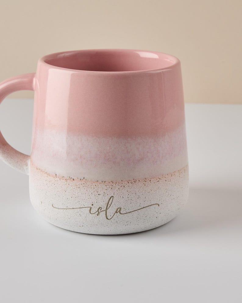May include: A pink and white ceramic mug with a textured finish. The mug has a white band around the middle and the word "isla" written in gold script on the bottom.