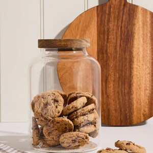 Peut inclure: Un bocal en verre transparent avec un couvercle en bois rempli de biscuits aux p&eacute;pites de chocolat. Le bocal est pos&eacute; sur une surface blanche &agrave; c&ocirc;t&eacute; d'une planche &agrave; d&eacute;couper en bois.