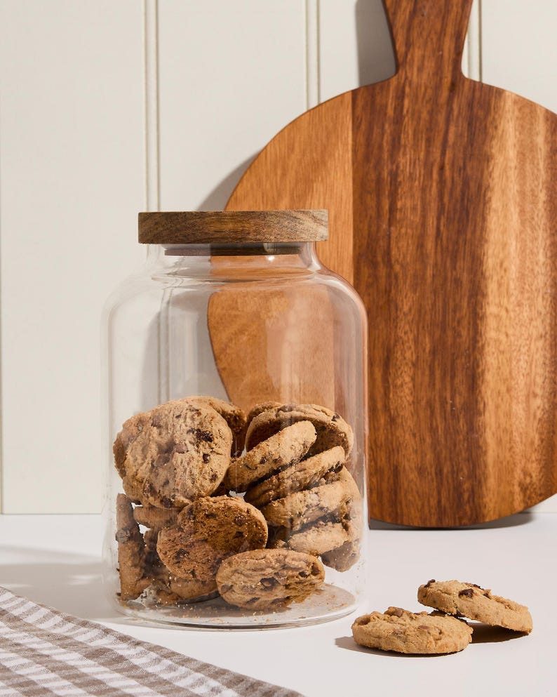 Peut inclure: Un bocal en verre transparent avec un couvercle en bois rempli de biscuits aux p&eacute;pites de chocolat. Le bocal est pos&eacute; sur une surface blanche &agrave; c&ocirc;t&eacute; d'une planche &agrave; d&eacute;couper en bois.