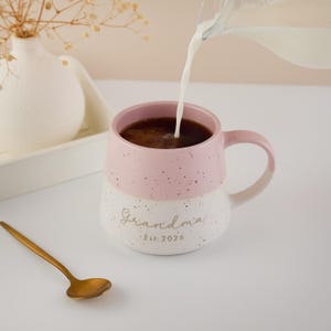 May include: A pink and white speckled ceramic mug with the word "Grandma" and "Est. 2026" printed on it. Coffee is being poured into the mug. A gold spoon rests on the table next to the mug.