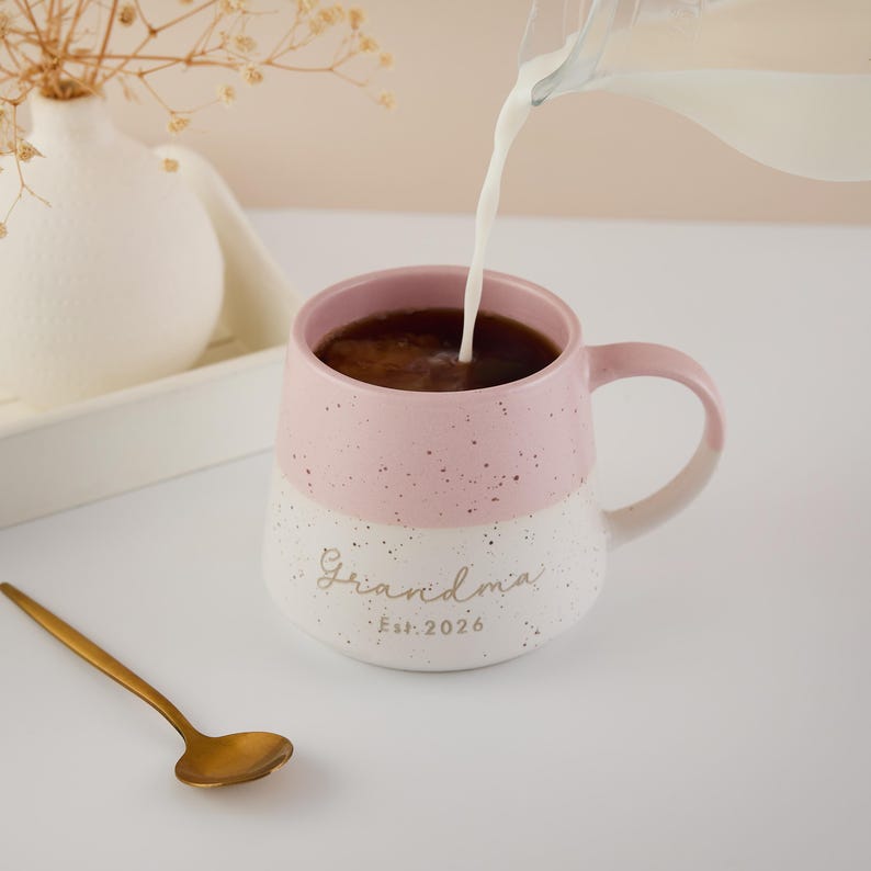 May include: A pink and white speckled ceramic mug with the word "Grandma" and "Est. 2026" printed on it. Coffee is being poured into the mug. A gold spoon rests on the table next to the mug.