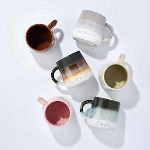 May include: Six ceramic mugs with different glaze patterns and personalized names. The mugs are in various colors, including brown, black, white, pink, and blue. The names on the mugs are "Ann", "Jackson", "Carl", and three other names that are not visible in the image.