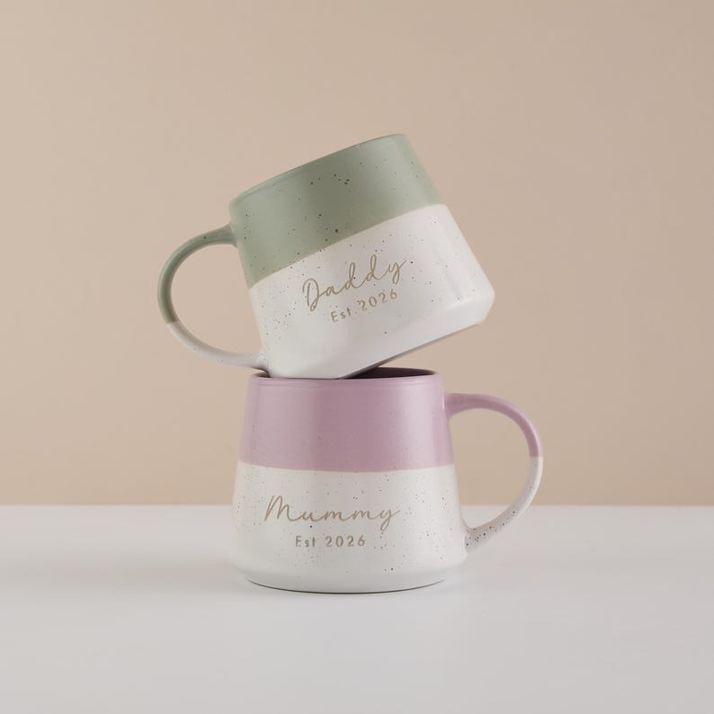 May include: Two ceramic mugs, one stacked on top of the other. The top mug is green and white, with the word "Daddy" and "Est 2026" printed on it. The bottom mug is pink and white, with the word "Mummy" and "Est 2026" printed on it.
