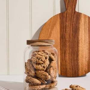 Peut inclure: Un bocal en verre avec un couvercle en bois rempli de biscuits aux p&eacute;pites de chocolat. Le bocal est pos&eacute; sur une surface blanche &agrave; c&ocirc;t&eacute; d'une planche &agrave; d&eacute;couper en bois.