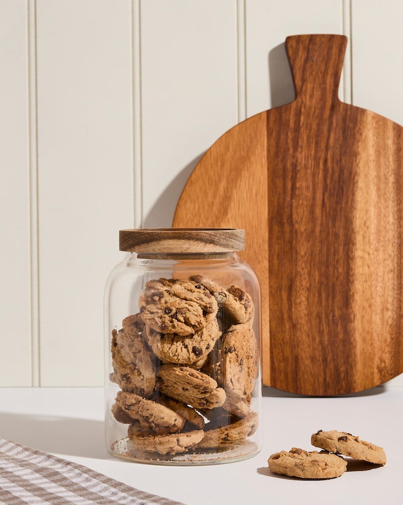 Peut inclure: Un bocal en verre avec un couvercle en bois rempli de biscuits aux p&eacute;pites de chocolat. Le bocal est pos&eacute; sur une surface blanche &agrave; c&ocirc;t&eacute; d'une planche &agrave; d&eacute;couper en bois.